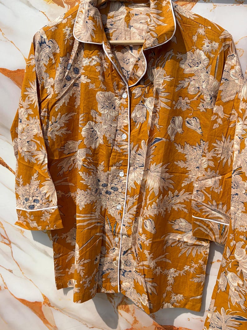 Pode incluir: Conjunto de pijama mostarda com um padr&atilde;o floral em creme e azul marinho. O conjunto inclui uma camisa com bot&otilde;es com debrum branco e cal&ccedil;as a condizer. O tecido parece leve e confort&aacute;vel.