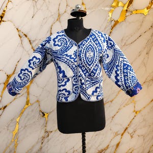 May include: A quilted jacket featuring a vibrant blue and white paisley pattern. This long-sleeve garment has a V-neckline, a front button closure, and two front pockets with piped edges. The fabric has a textured, grid-like surface finish.