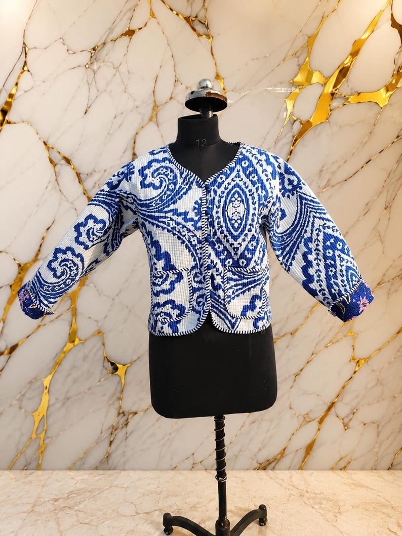 May include: A quilted jacket featuring a vibrant blue and white paisley pattern. This long-sleeve garment has a V-neckline, a front button closure, and two front pockets with piped edges. The fabric has a textured, grid-like surface finish.