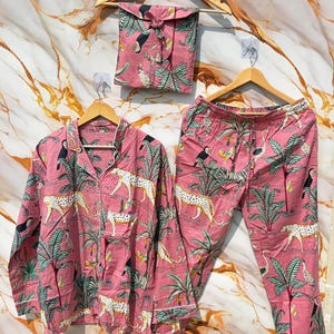 Może przedstawiać: R&oacute;żowy zestaw piżamowy z tropikalnym nadrukiem z tukanami, lampartami i palmami. Zestaw zawiera koszulę zapinaną na guziki, spodnie i pasującą torbę ze sznurkiem, wszystko zawieszone na drewnianych wieszakach.