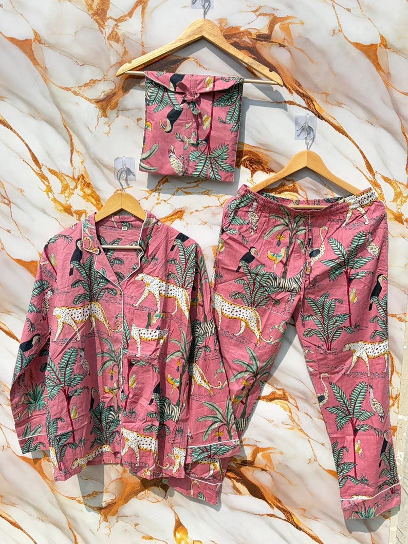 Może przedstawiać: R&oacute;żowy zestaw piżamowy z tropikalnym nadrukiem z tukanami, lampartami i palmami. Zestaw zawiera koszulę zapinaną na guziki, spodnie i pasującą torbę ze sznurkiem, wszystko zawieszone na drewnianych wieszakach.