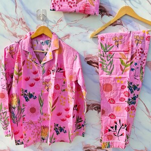 May include: Pink floral pajama set, comprising a button-down shirt, trousers, and a matching pouch. The floral pattern includes red, yellow, and blue flowers with green leaves. The pajamas are displayed on wooden hangers.