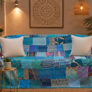 May include: A blue patchwork quilt draped over a sofa, featuring various shades of teal, navy, and turquoise with intricate kantha stitching. Two solid white square throw pillows are placed on top of the quilt, which displays a mix of floral, geometric, and abstract patterns across its rectangular fabric panels.