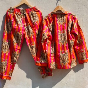 K&ouml;nnte beinhalten: Ein zweiteiliges Pyjama-Set mit Tigerstreifenmuster in Orange, Pink und Beige. Das lang&auml;rmlige Oberteil und die Hose werden auf Holzb&uuml;geln pr&auml;sentiert. Wei&szlig;er Besatz an den Kanten, vor neutralem Hintergrund.
