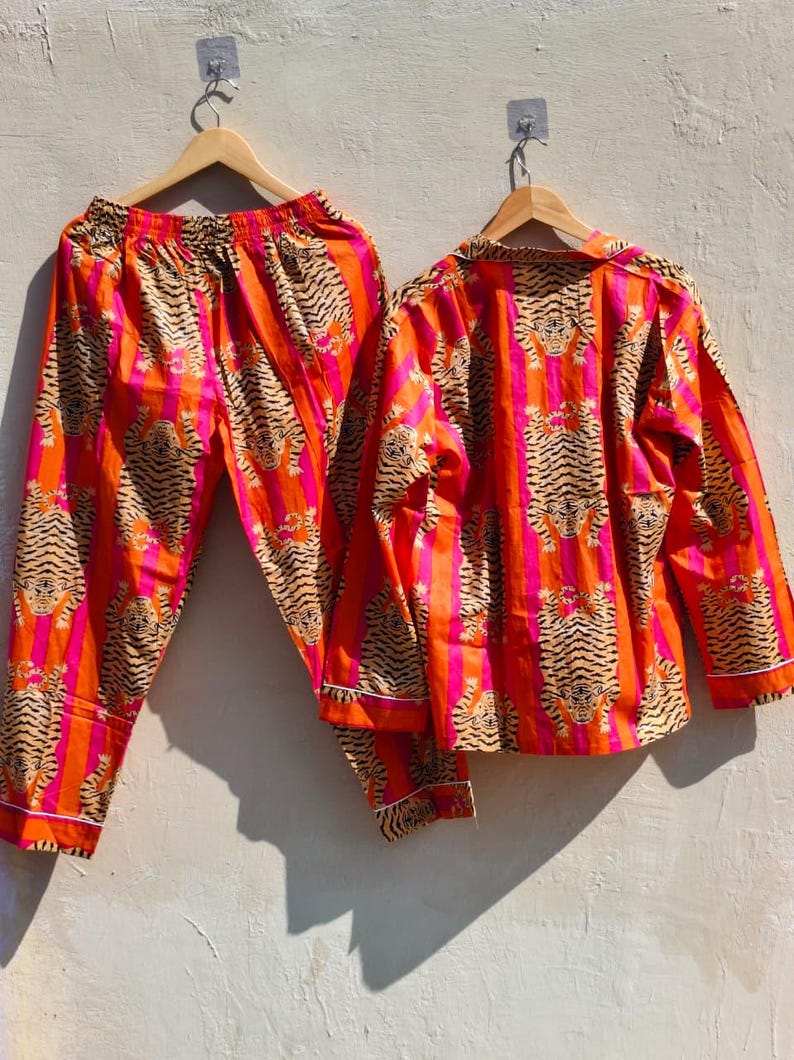 K&ouml;nnte beinhalten: Ein zweiteiliges Pyjama-Set mit Tigerstreifenmuster in Orange, Pink und Beige. Das lang&auml;rmlige Oberteil und die Hose werden auf Holzb&uuml;geln pr&auml;sentiert. Wei&szlig;er Besatz an den Kanten, vor neutralem Hintergrund.