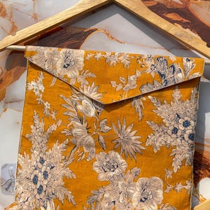 Pode incluir: Tecido amarelo mostarda com um padr&atilde;o floral em branco e azul marinho. O tecido &eacute; dobrado em forma quadrada e pendurado em um cabide de madeira. O design floral apresenta grandes flores e folhagens.