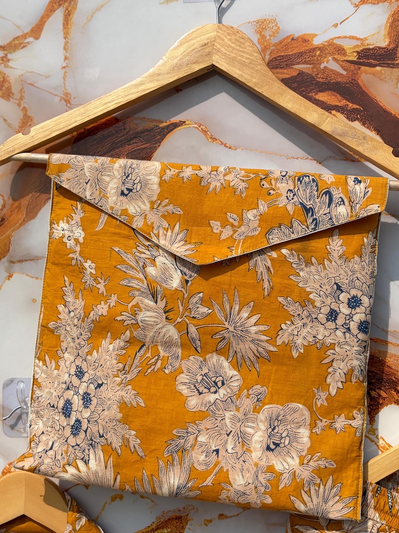 Pode incluir: Tecido amarelo mostarda com um padr&atilde;o floral em branco e azul marinho. O tecido &eacute; dobrado em forma quadrada e pendurado em um cabide de madeira. O design floral apresenta grandes flores e folhagens.