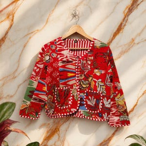 May include: A red quilted jacket with a vibrant floral and geometric pattern. The jacket features a round neckline, long sleeves, and two front pockets. The jacket is hanging on a wooden hanger against a marble background.