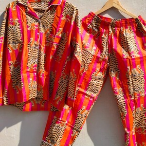 K&ouml;nnte beinhalten: Ein zweiteiliges Pyjama-Set mit einem Tigerstreifenmuster in Orange, Pink und Schwarz. Das lang&auml;rmlige Hemd und die passende Hose werden auf Holzb&uuml;geln pr&auml;sentiert. Der Pyjama hat eine wei&szlig;e Paspelierung.
