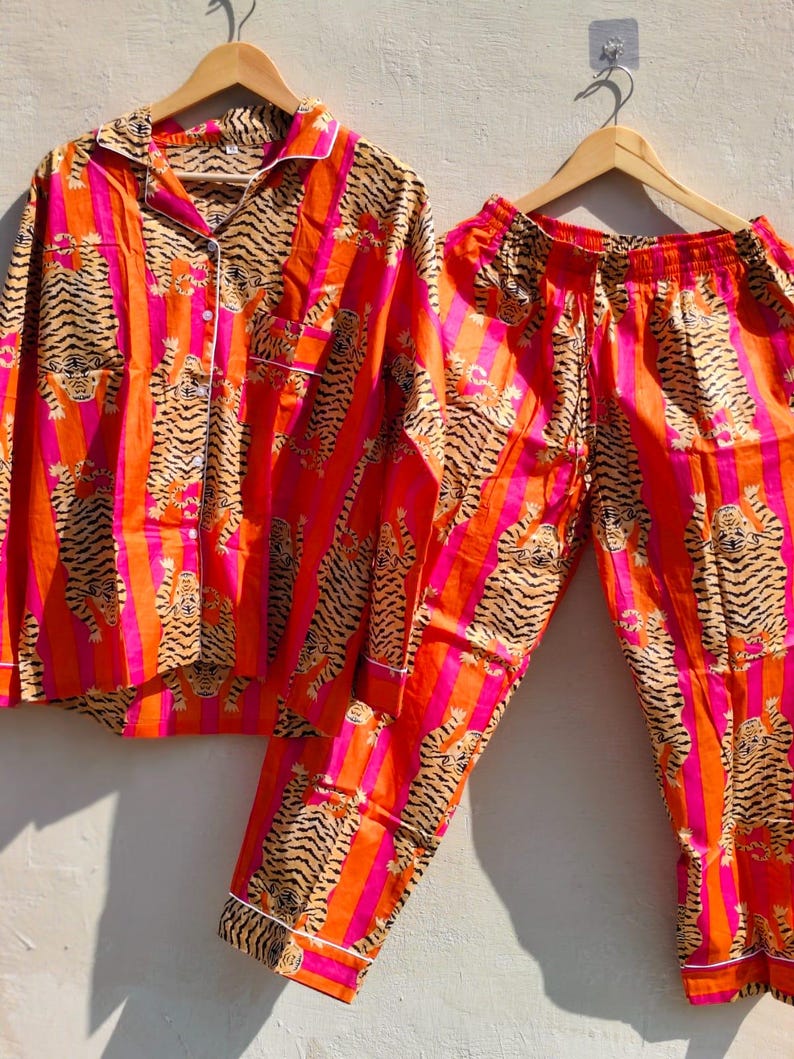 K&ouml;nnte beinhalten: Ein zweiteiliges Pyjama-Set mit einem Tigerstreifenmuster in Orange, Pink und Schwarz. Das lang&auml;rmlige Hemd und die passende Hose werden auf Holzb&uuml;geln pr&auml;sentiert. Der Pyjama hat eine wei&szlig;e Paspelierung.