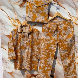 Pode incluir: Conjunto de pijama mostarda com padr&atilde;o floral. O conjunto inclui uma camisa com bot&otilde;es, cal&ccedil;as com cord&atilde;o e uma bolsa a condizer. As bordas brancas acentuam a gola, os punhos e a bainha das cal&ccedil;as. Os pijamas s&atilde;o exibidos em cabides de madeira.