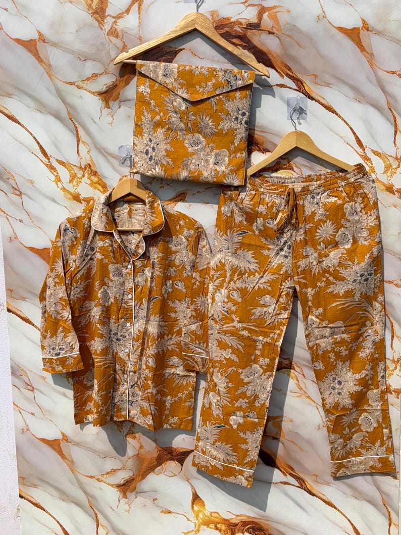 Pode incluir: Conjunto de pijama mostarda com padr&atilde;o floral. O conjunto inclui uma camisa com bot&otilde;es, cal&ccedil;as com cord&atilde;o e uma bolsa a condizer. As bordas brancas acentuam a gola, os punhos e a bainha das cal&ccedil;as. Os pijamas s&atilde;o exibidos em cabides de madeira.