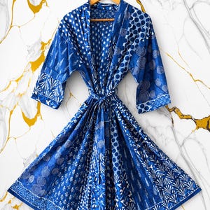 May include: A blue and white patterned robe with a tie waist. The robe features a variety of geometric and floral designs in shades of blue and white. The robe is hanging on a wooden hanger against a marble background.