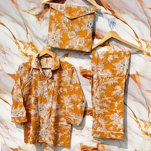 Pode incluir: Conjunto de pijama mostarda com padr&atilde;o floral, incluindo uma camisa com bot&otilde;es, cal&ccedil;as e uma bolsa de armazenamento a condizer. O conjunto &eacute; exibido em cabides de madeira sobre um fundo com padr&atilde;o de m&aacute;rmore.