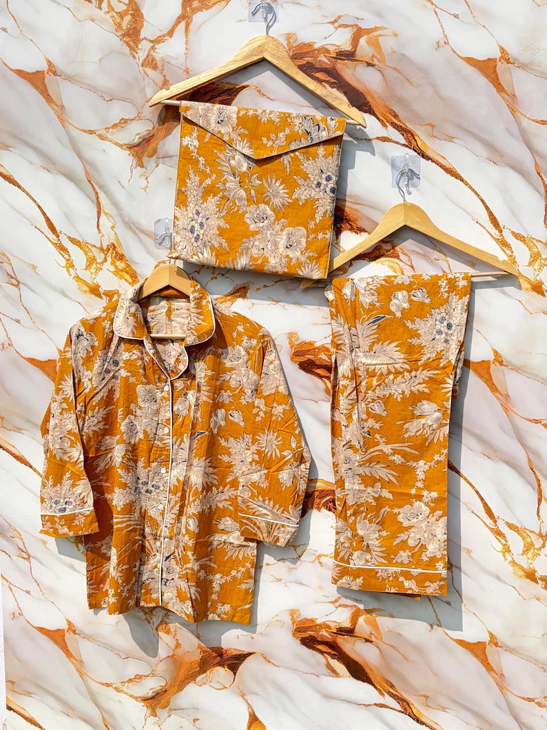 Pode incluir: Conjunto de pijama mostarda com padr&atilde;o floral, incluindo uma camisa com bot&otilde;es, cal&ccedil;as e uma bolsa de armazenamento a condizer. O conjunto &eacute; exibido em cabides de madeira sobre um fundo com padr&atilde;o de m&aacute;rmore.