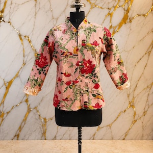 May include: A light pink quilted jacket featuring a mandarin collar, long sleeves with folded cuffs, and a front button closure. The fabric is decorated with a vibrant floral and bird pattern in shades of red, green, yellow, and brown.