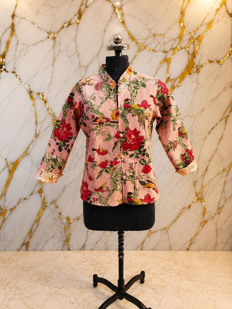 May include: A light pink quilted jacket featuring a mandarin collar, long sleeves with folded cuffs, and a front button closure. The fabric is decorated with a vibrant floral and bird pattern in shades of red, green, yellow, and brown.