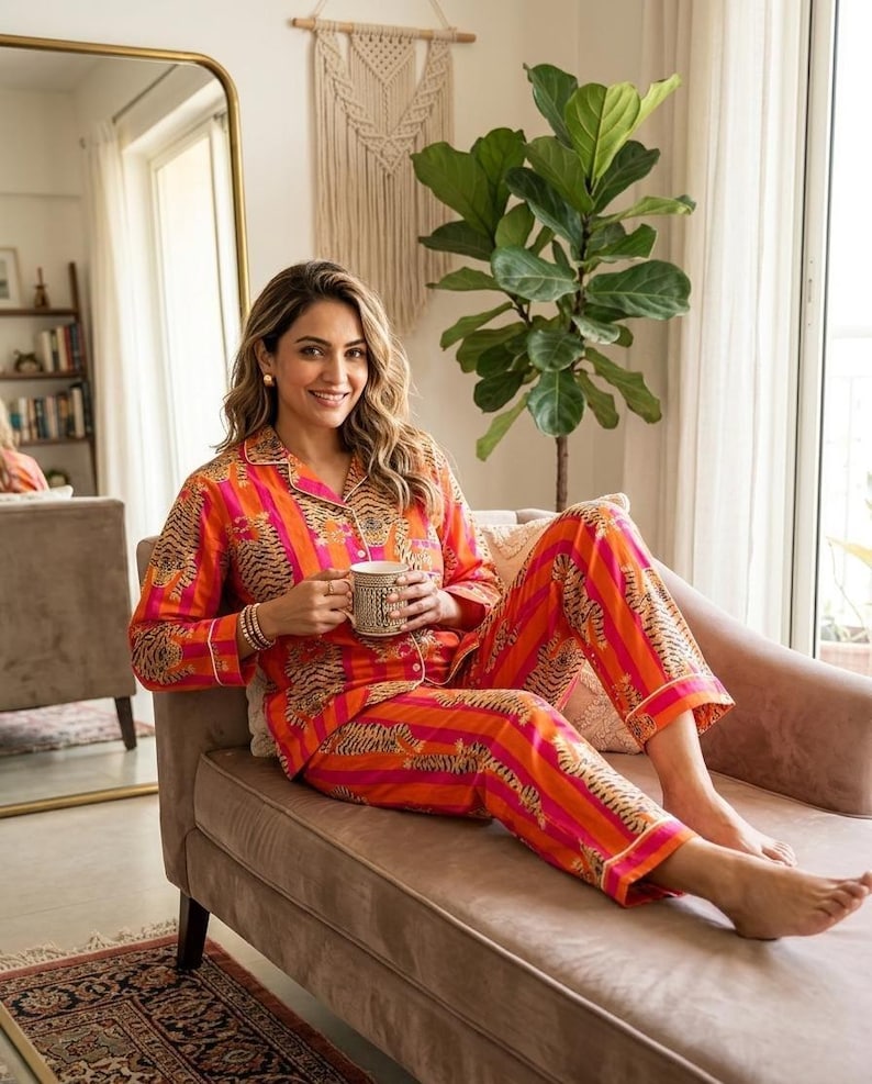 K&ouml;nnte beinhalten: Ein orange-pinkfarbener Pyjama-Set mit Tigerstreifenmuster. Das lang&auml;rmlige Oberteil und die Hose werden von einer Person getragen, die auf einem braunen Sofa sitzt und eine Tasse h&auml;lt. Der Pyjama hat wei&szlig;e Paspeln.