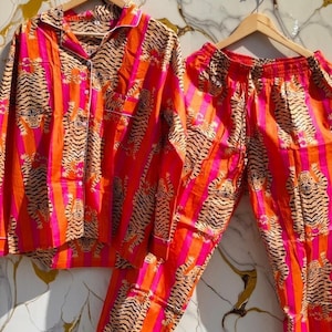 K&ouml;nnte beinhalten: Ein lebendiges Pyjama-Set mit Tigerstreifenmuster in Orange, Pink und Schwarz. Das Set beinhaltet ein Hemd mit Kn&ouml;pfen und Brusttasche sowie eine passende Hose. Wei&szlig;e Paspeln akzentuieren Kragen, Manschetten und Tasche.