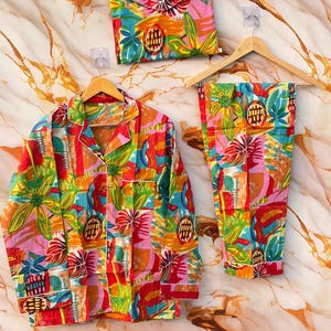 May include: A bright, patterned pajama set featuring a floral and abstract design. The set includes a shirt, trousers, and a matching pouch, displayed on wooden hangers. The colour scheme is a mix of red, orange, green, and blue.