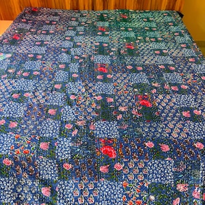 May include: A rectangular quilt featuring a patchwork design of various blue, green, and white floral patterns. The fabric is covered in small, repeating motifs of pink and red flowers, with visible running stitch quilting throughout the entire surface.