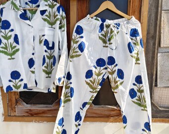 May include: White pajama set with a blue floral pattern, including a button-down shirt, drawstring pants, and a matching pouch. The floral design features blue flowers and green leaves. The set is displayed on wooden hangers.