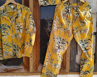 May include: Yellow pajama set with a tropical print featuring palm trees and leopards. The set includes a button-down shirt, pants, and a matching eye mask, all hanging on wooden hangers. The fabric has a white trim.