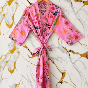 May include: A pink floral print robe with a tie closure. The robe features a vibrant pattern of flowers and leaves in shades of pink, yellow, blue, and green. The robe is hanging on a wooden hanger.