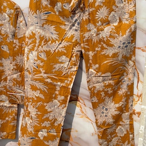 Pode incluir: Conjunto de pijama mostarda com estampa floral em branco e azul. O conjunto inclui uma parte superior de manga comprida e cal&ccedil;as com cintura com cord&atilde;o e acabamento branco. O tecido tem uma apar&ecirc;ncia macia e fluida.