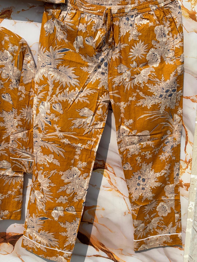 Pode incluir: Conjunto de pijama mostarda com estampa floral em branco e azul. O conjunto inclui uma parte superior de manga comprida e cal&ccedil;as com cintura com cord&atilde;o e acabamento branco. O tecido tem uma apar&ecirc;ncia macia e fluida.