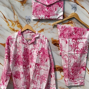 Puede incluir: Un conjunto de pijama rosa y blanco con una bolsa de almacenamiento a juego con forma de sobre. El conjunto incluye una camisa con botones, pantalones y una bolsa, todos con un estampado tropical con árboles, pájaros y tigres. El conjunto se exhibe en perchas de madera.