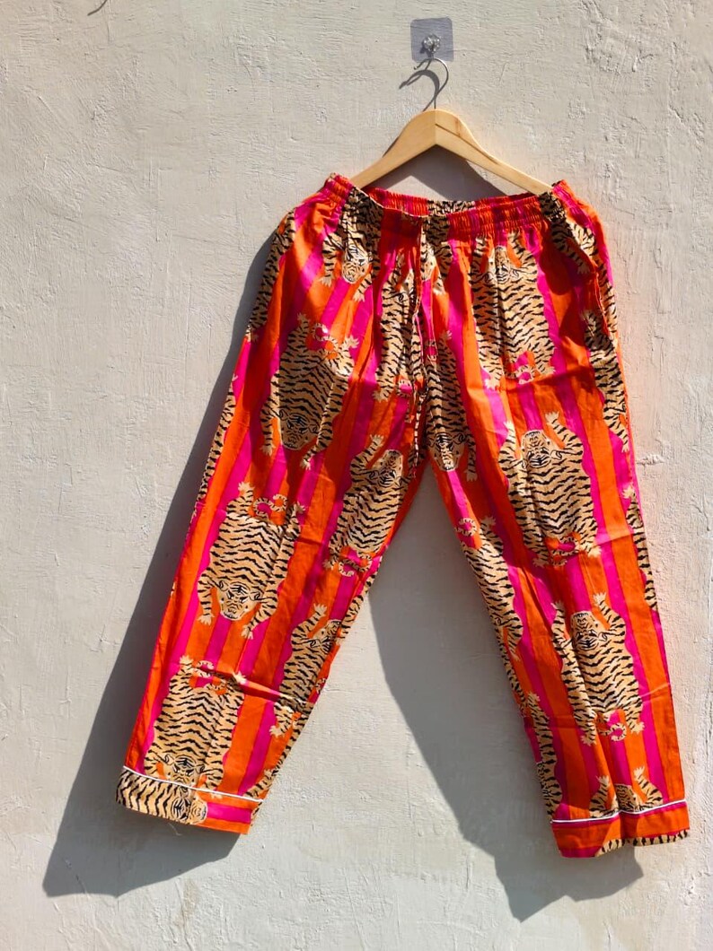 K&ouml;nnte beinhalten: Orangefarbene und pinkfarbene Schlafanzughose mit Tigerstreifenmuster. Die Hose hat einen Kordelzug in der Taille und wei&szlig;e Paspeln an den Manschetten. Die Tigerstreifen sind in einem beigen Farbton gehalten.