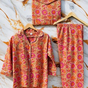 Op de afbeelding: Een pyjamaset bestaande uit een shirt met lange mouwen en knoopsluiting en een bijpassende broek, vergezeld van een rechthoekig tasje in envelopstijl. De stof heeft een levendig bloemenpatroon in tinten felroze, oranje en perzik op een lichte achtergrond.