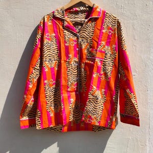 K&ouml;nnte beinhalten: Ein lang&auml;rmeliges Hemd mit Tigerstreifenmuster in Orange, Pink und Schwarz. Das Hemd hat eine Knopfleiste und wei&szlig;e Paspeln. Es h&auml;ngt an einem Holzb&uuml;gel. Die Ma&szlig;e sind in Zentimetern.