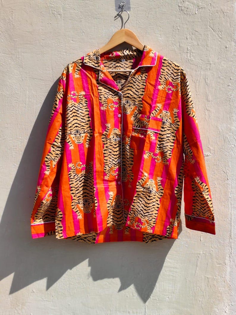 K&ouml;nnte beinhalten: Ein lang&auml;rmeliges Hemd mit Tigerstreifenmuster in Orange, Pink und Schwarz. Das Hemd hat eine Knopfleiste und wei&szlig;e Paspeln. Es h&auml;ngt an einem Holzb&uuml;gel. Die Ma&szlig;e sind in Zentimetern.