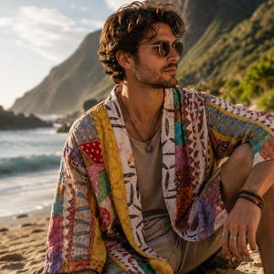 May include: A colorful patchwork kimono-style jacket with geometric and floral patterns. The jacket features a mix of red, yellow, purple, and blue fabrics. The person is wearing sunglasses and a necklace, sitting on a beach with mountains in the background.
