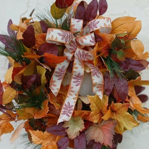 May include: A fall-themed wreath composed of colorful artificial leaves in shades of orange, yellow, and burgundy. A white ribbon with a leaf pattern is tied in a bow at the top. Green pine needles are interspersed throughout the leaves.