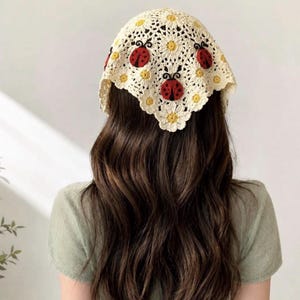 Peut inclure: Un foulard triangulaire en crochet de couleur crème présentant un motif de marguerites répétitif avec des centres jaunes. L'accessoire est orné de trois appliqués de coccinelles rouges et noires cousus sur le tissu floral.