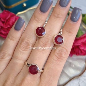 Könnte beinhalten: Ein Schmuckset mit einem Ring und einem Paar Ohrringen, jeweils mit einem großen, runden, tiefroten Edelstein. Der Ring und die Ohrringe sind in Silbermetall gefasst. Der Text "jewelrycomplex" ist sichtbar.
