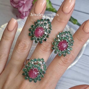 Puede incluir: Un conjunto de joyas de tres piezas con un anillo y dos pendientes. Cada pieza tiene una gema rosa ovalada central rodeada de pequeñas gemas verdes. Las joyas se presentan sobre un fondo claro. El texto "Jewelrycomplex" es visible.