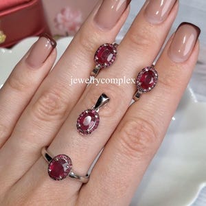 Puede incluir: Un conjunto de joyas con un anillo, un colgante y dos pendientes. Cada pieza tiene un engaste plateado y una gema roja ovalada grande rodeada de piedras rojas más pequeñas. El texto "jewelrycomplex" es visible.