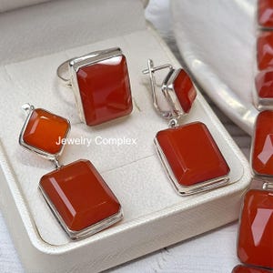 May include: A jewelry set featuring a silver ring and two drop earrings, each set with rectangular, faceted orange-red carnelian stones. The ring has a single stone, while the earrings feature two stones each. The text Jewelry Complex is visible.