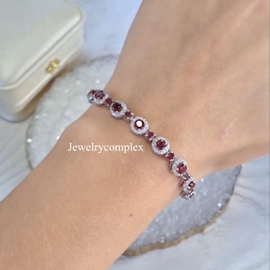 May include: A silver bracelet featuring a sequence of circular, deep red gemstones, each encircled by smaller, clear stones. The bracelet is shown on a wrist, with the text "Jewelrycomplex" visible.
