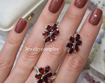 May include: A jewelry set featuring a ring and earrings, each designed with a flower motif. The petals are made of dark red gemstones, set in a silver metal. The text "Jewelrycomplex" is visible in the image.
