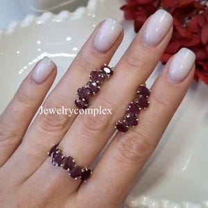 May include: A silver ring and two matching silver earrings, each featuring a row of five oval-shaped deep red gemstones set in metal prongs. The text Jewelrycomplex is visible in the centre of the image, positioned between the ring and the earrings.