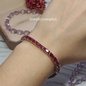 May include: A silver-toned tennis bracelet featuring a continuous row of oval-cut, deep red gemstones set in metal prongs. The text Jewelrycomplex is visible in the centre of the frame, positioned above the bracelet on the skin.