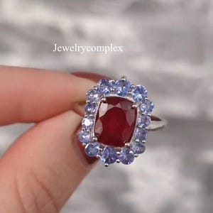 Könnte beinhalten: Ein silberner Ring mit einem großen, rechteckigen, tiefroten Edelstein, umgeben von kleineren, hellblauen Edelsteinen. Der Ring ist vor einem verschwommenen Hintergrund platziert. Der Text "jewelrycomplex" ist sichtbar.