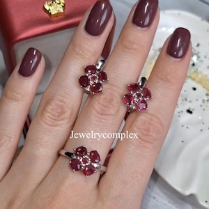 Puede incluir: Un conjunto de anillo y pendientes con un diseño floral con piedras preciosas rojas oscuras de corte ovalado. El anillo está en un dedo y los pendientes se muestran junto a él. La joyería se coloca sobre un fondo neutro.