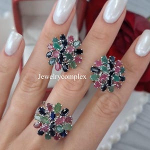 Op de afbeelding: Een set van drie ringen met een bloemmotief. Elke ring heeft een cluster van kleurrijke edelstenen, waaronder groene, roze, blauwe en zwarte stenen, gezet in een zilverkleurig metaal. De ringen worden getoond op een hand met witte nagellak.