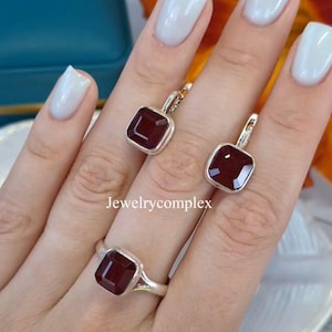 Puede incluir: Un conjunto de joyas con un anillo y un par de pendientes, cada uno con una piedra preciosa cuadrada de color rojo intenso. El anillo y los pendientes tienen un engaste de metal plateado. El texto "Jewelrycomplex" es visible.